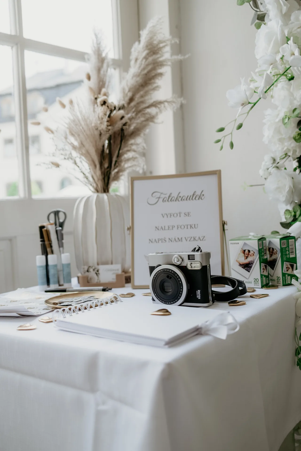 Stolek fotokoutku s cedulí, fotoaparátem Instax a knihou hostů vedle bílé květinové stěny